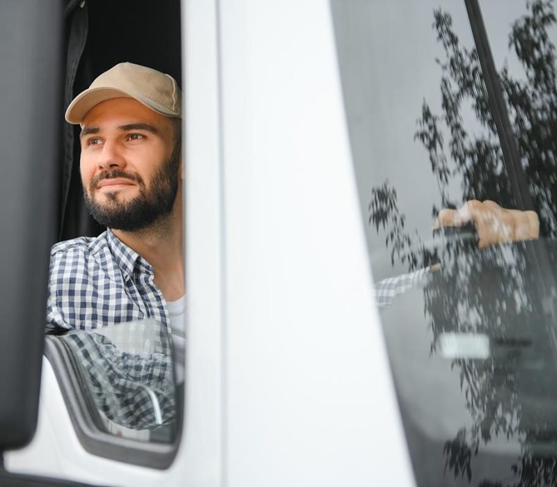 A driver sitting inside the semi-truck while looking through the open window with multiple trucks parked in the background.